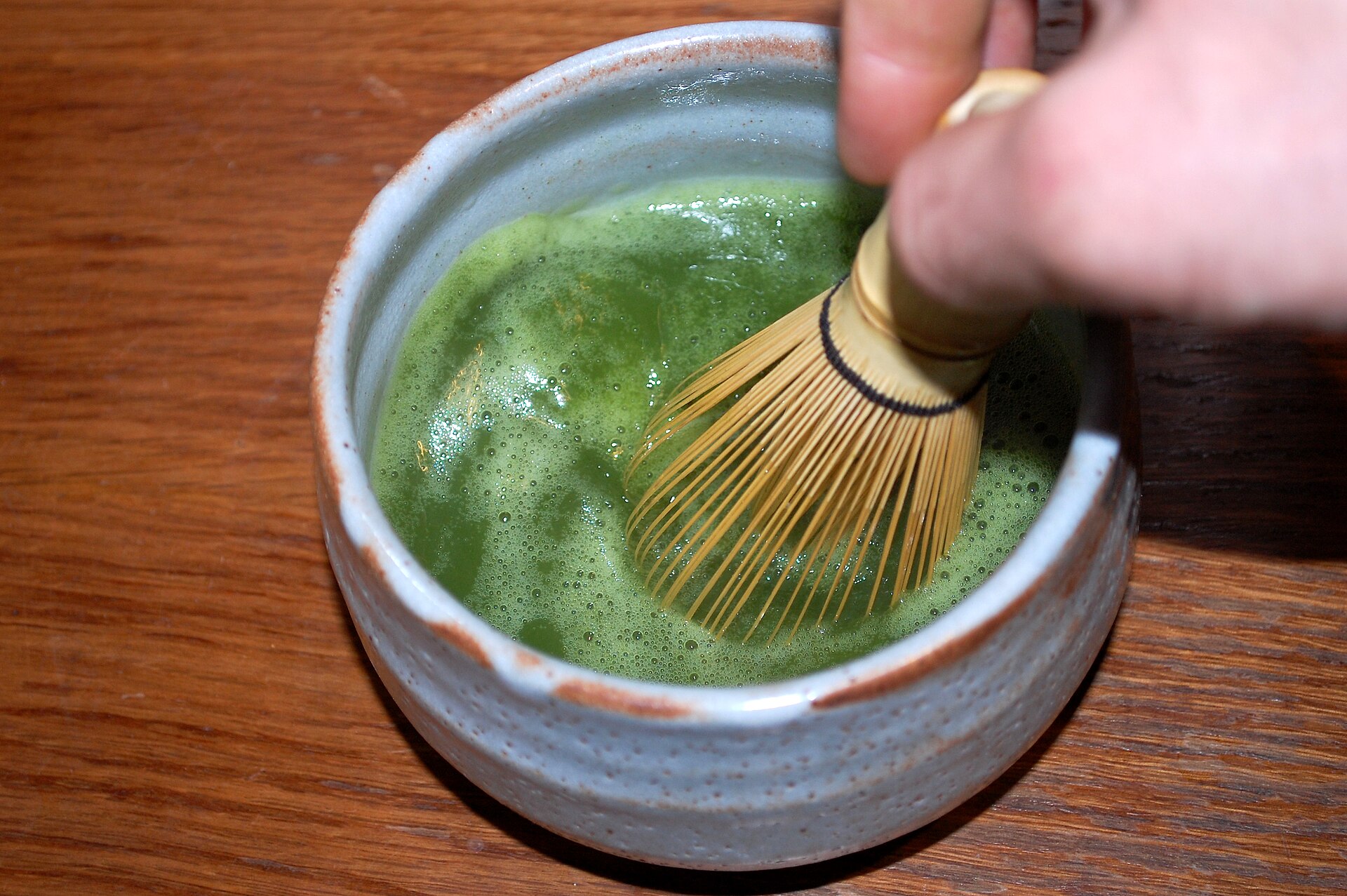Real photograph of a hand whisking bright green matcha with a bamboo whisk in a ceramic bowl